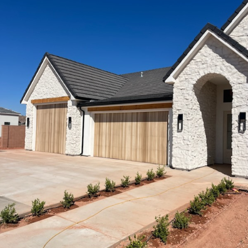 Three-car garage with Vidor wood overlay garage doors in a natural finish, installed on a modern white brick home with black roofing in Southern Utah.